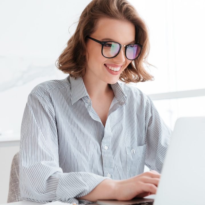 Close-up portrait of cheerful bunette girl in glasses using laptop computer while working at home